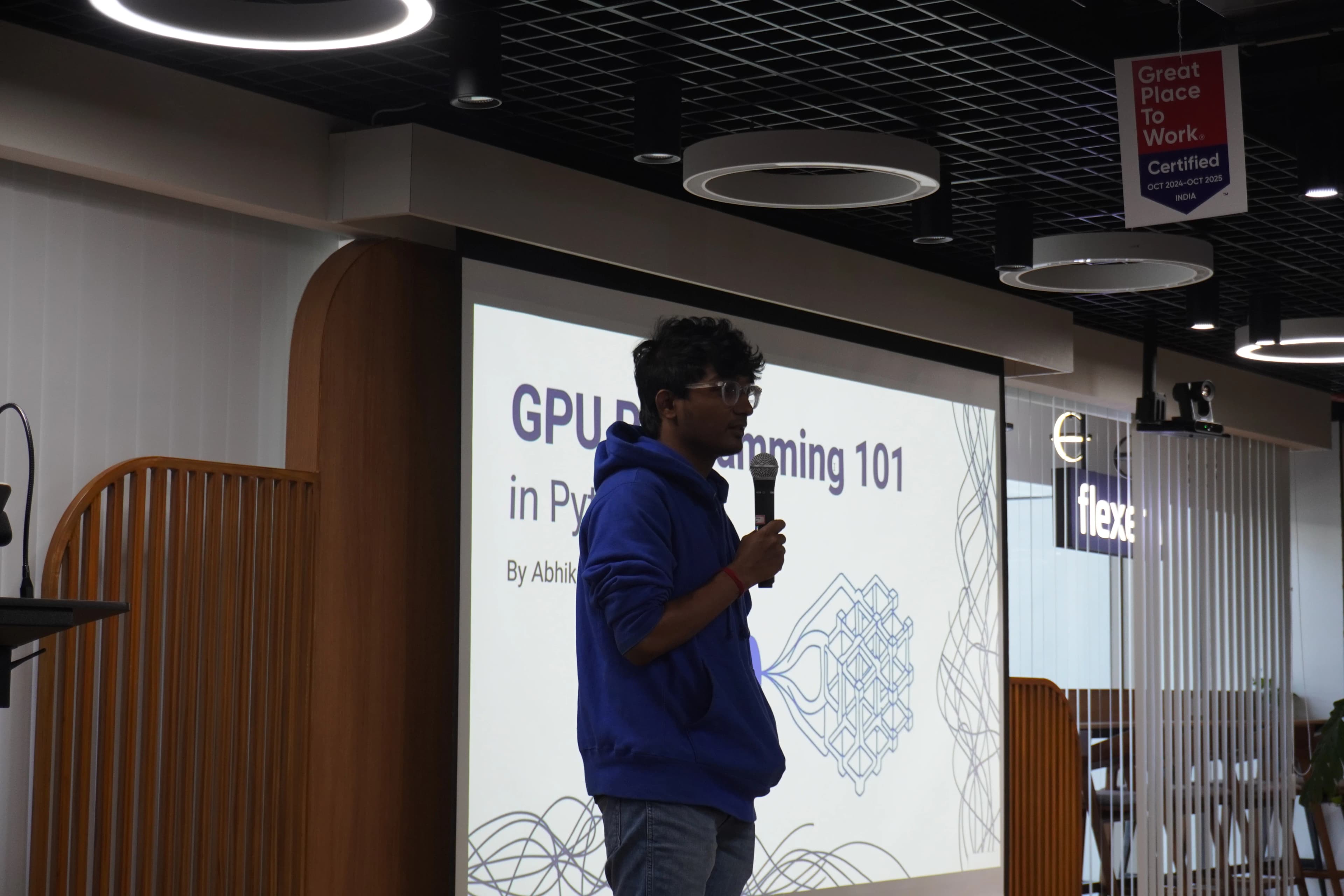 Abhik Sarkar presenting GPU Programming 101 in Python at the BangPypers December 2025 meetup, standing with a microphone in front of a projected slide showing the talk title and a geometric GPU illustration, at the Flexera office in Bengaluru