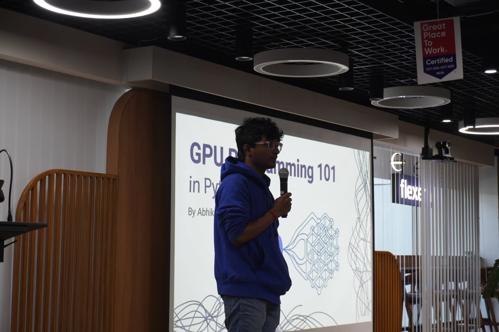 Abhik Sarkar presenting GPU Programming 101 in Python at the BangPypers December 2025 meetup, standing with a microphone in front of a projected slide showing the talk title and a geometric GPU illustration, at the Flexera office in Bengaluru