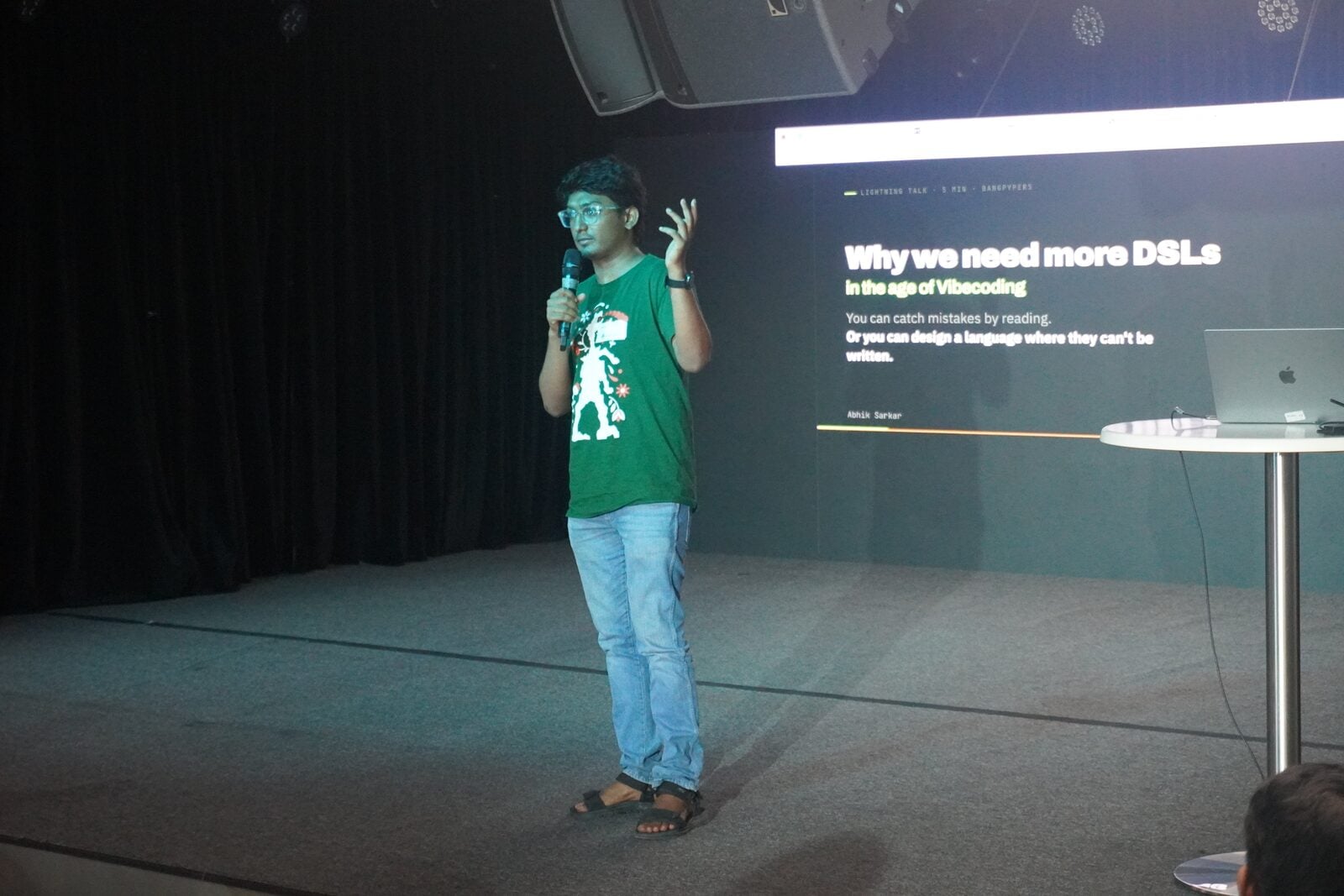 Abhik Sarkar giving a lightning talk on stage with a microphone, beside a projected slide titled "Why we need more DSLs in the age of Vibecoding" at import_ bengaluru 2026
